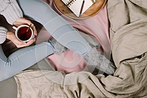 Cozy flatlay of woman`s legs in blue leggins and warm grey socks in bed with cup of tea