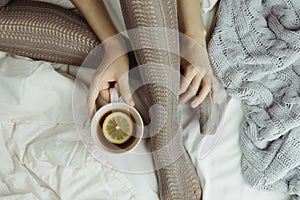 Cozy flatlay of woman`s legs in warm grey stockings in bed with knitted sweater and blanket aside