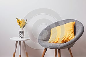 Cozy grey chair with yekllow pillow and flowers in the vase standing near the white wall.