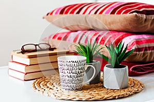 Cozy home interior decor: cup of coffee, stack of books, plants in pots on a wicker stand, pillows and plaid on a white table.