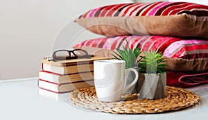 Cozy home interior decor: cup of coffee, stack of books, plants in pots on a wicker stand, pillows and plaid on a white table.