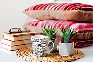 Cozy home interior decor: cup of coffee, stack of books, plants in pots on a wicker stand, pillows and plaid on a white table.