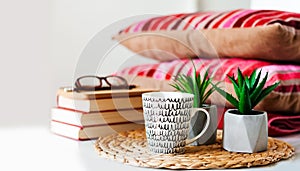 Cozy home interior decor: cup of coffee, stack of books, plants in pots on a wicker stand, pillows and plaid on a white table.