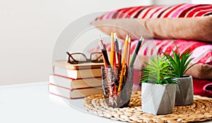 Cozy home interior decor: pencils in holder, stack of books, plants in pots on a wicker stand, pillows and plaid on a white table