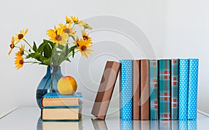 Cozy home interior decor: stack of books, peach and vase with yellow flowers on a table. Distance home education.Quarantine