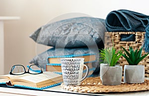 Cozy home interior decor: stack of books, plants in pots on a wicker stand, pillows and plaid on a white table. Distance home