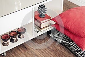 Cozy home interior decor, white nightstand with pile of books, candles in coconut shell with decorative pillows background