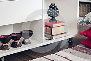 Cozy home interior decor, white nightstand with pile of books, candles in coconut shell near a multi-colored rug with decorative