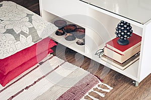 Cozy home interior decor, white nightstand with pile of books, candles in coconut shell near a multi-colored rug with decorative