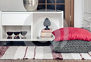 Cozy home interior decor, white nightstand with pile of books, candles in coconut shell near a multi-colored rug with decorative