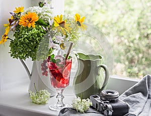 Cozy home still life - bouquet of flowers, glass of lemonade, jug, vintage camera on the window in a bright room