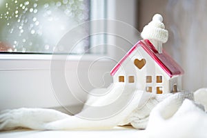 Cozy house is wrapped in a hat and scarf in a snowstorm -window sill decor. Winter, snow - home insulation, protection from cold