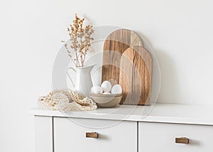 Cozy kitchen interior. Dishes, cutting boards, bowl with eggs, a jug of dried flowers on a white cabinet