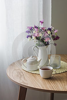 Cozy kitchen interior - a jug with summer garden flowers, a cup of tea, a teapot on a round wooden table