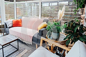 Cozy, light conservatory interior design with gray sofa decorated with bright textile cover and cushions and many green