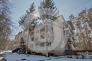 Cozy old three-storey concrete multifamily apartment building in a coniferous forest on a sunny winter day