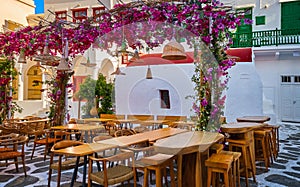 Cozy outdoor dining, cafe, restaurant, taverna in small Greek square, whitewashed chapel, vibrant pink bougainvillea in