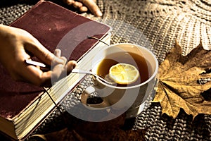 Cozy photo of female hands, a cup of tea with lemon on the background of knitted sweater, yellow dry autumn leaves and an old book