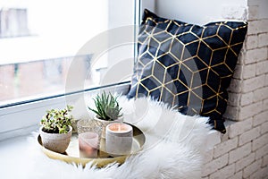 Cozy real home decoration, burning candles on golden tray with pillow on white faux fur on windowsill