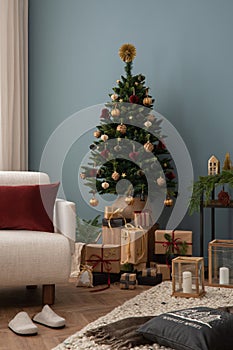 Cozy and stylish christmas living room interior with design armchair, christmas tree, gifts, wooden laterns, blue wall, red pillow