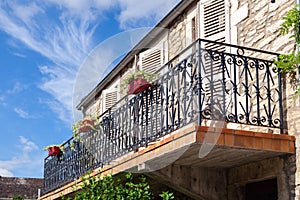 Cozy vintage French balcony with black metal railings, flowers in pot, open shutters on windows against blue sky, clouds. Bottom
