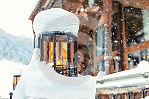 Cozy vintage street lamp with orange glass covered by snow and wooden rustic house on background outdoors at snowfall. Retro metal