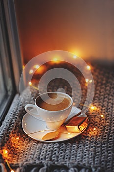 Cozy winter or autumn morning at home. Hot coffee with gold metallic spoon, warm blanket, garland and candle lights