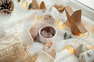 Cozy winter. Hands in sweater holding stylish cup of tea with modern christmas decoration, pine cone, wooden star and tree, golden