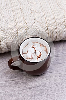 Cozy winter home background, cup of hot cocoa with marshmallow, warm knitted sweater on white wooden board background.