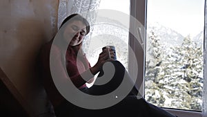 Cozy winter lifestyle. Young happy woman drinking cup of coffee wearing knitted sweater sitting home by the big window
