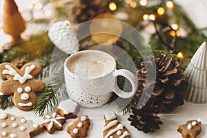Cozy winter mood. Coffee with gingerbread cookies, pine cones and warm lights on white wooden table