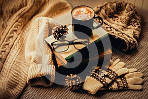 A cozy winter still life, featuring warm woolen clothing, books, glasses, pinecones, and a steaming cup of coffee with a hint of