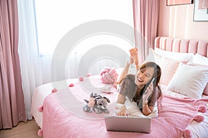 Cozy young asian woman freelancer laying on modern bed and typing laptop with lamp in bed room at home