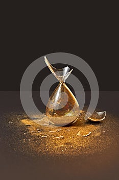 Cracked antique hourglass with golden sand on a black duotone background.