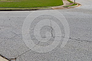 Cracked asphalt intersection with formed curbs and green grass border, aged and weathered street surface