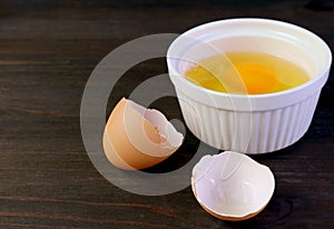 Cracked eggshell on dark brown wooden table with blurry raw egg bowl in background