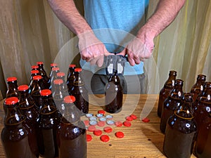 Craft beer brewing at home, man closes brown glass beer bottles with plastic capper on wooden table with red crown caps.