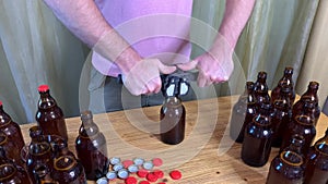 Craft beer brewing at home, man closes brown glass beer bottles with plastic capper on wooden table with red crown caps. Close up