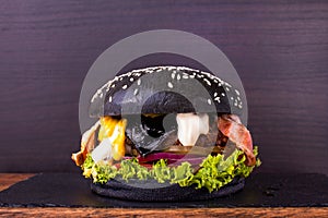 Craft black beef burger with black cheese, bacon, vegetables, onion jam, arugula on a board with a stone and wooden deck