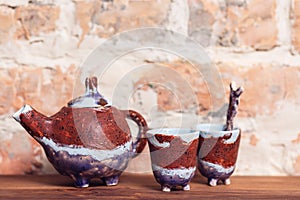 Craft ceramic teapot and cup for tea ceremony standing on old wooden shelf against brick wall