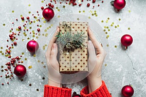 Craft gift of brown paper with twigs of Christmas tree and twine in the hands of a girl in a red sweater. Handmade gift on a gray