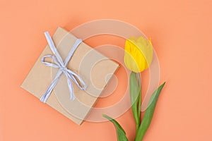 Craft paper gift box with white ribbon and single yellow tulip on an orange background