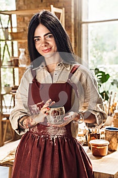 Craftsperson Concept. Young woman artist in twig wreath standingnear table with beautiful handmade cups smiling