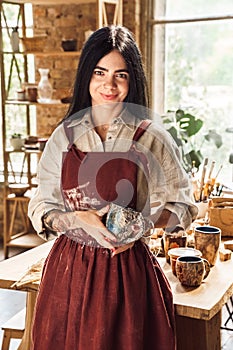 Craftsperson Concept. Young woman artist in twig wreath standingnear table with beautiful handmade cups smiling