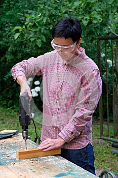 Craftswoman with safety glasses drilling a hole with an electric drill machine in wooden plank. Woodwork, DIY, gender equality