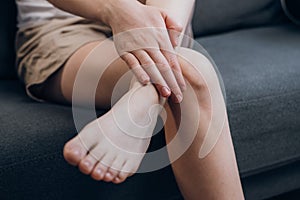 Cramps in leg calves or sprain calf on young caucasian female sitting on couch at home. Unhealthy woman with sprain is touching
