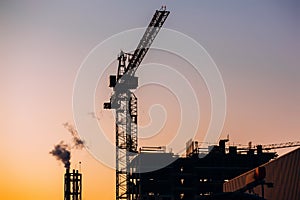 Crane and building construction site with pipe with smoke on background of sunset sky. Industrial landscape with silhouettes of c