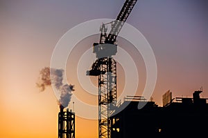 Crane and building construction site with pipe with smoke on background of sunset sky. Industrial landscape with silhouettes of c