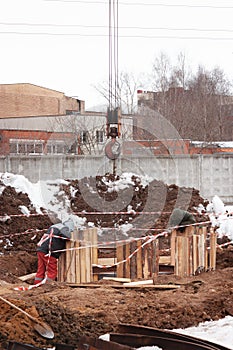 A crane hook going down into a pit, surrounded with wooden palette. The hook is attached on the wire going up to the frame. 2 men