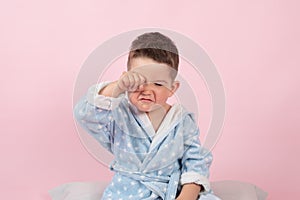 Cranky child boy refuses to go to bed. Kid boy in bathrobe on pink background